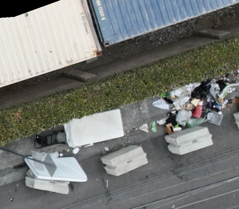 Bulky discarded items and mixed waste beside concrete barriers and shipping containers